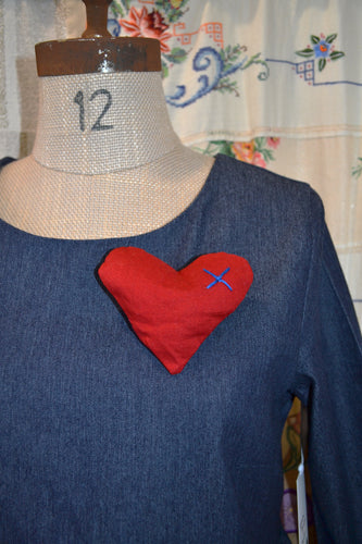 Large Red cotton brooch in the shape of a heart. It is stuffed to fill it out to a  3D form. It has a blue cross embroidered on the top left side of the heart. The brooch is pinned to a denim dress on a vintage mannequin.