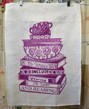 Load image into Gallery viewer, A white tea towel is clipped to a wooden hanger. there is a print in purple ink on the tea towel whihc shows a  stack of books with cups and plates on top. On the spine of the books reads &#39;My hobbies include ignoring the dishes and reading&quot;